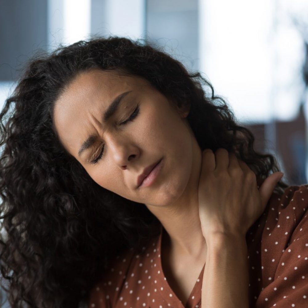 Close-up photo of curly-haired woman at home near the window with severe neck pain, mulatto business woman massaging her neck with closed eyes