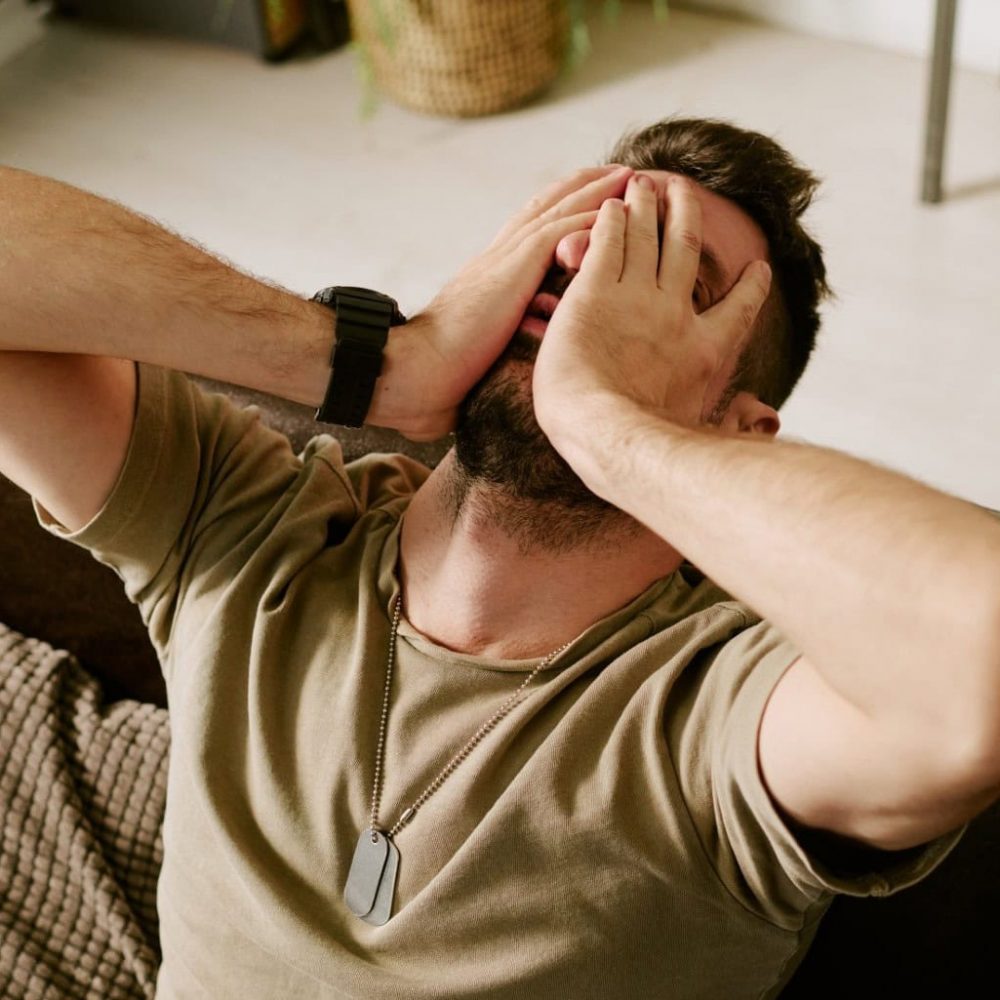 Middle aged Caucasian man sitting on couch covering face with hands showing signs of emotional distress representing psychological struggle or mental health issue
