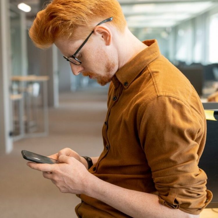 Young serious businessman in eyeglasses texting in smartphone or scrolling through contacts against his colleague standing by glass wall